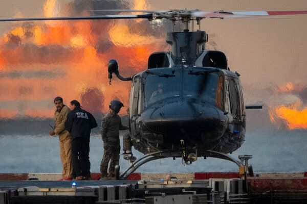 Un helicóptero oscuro está sobre una plataforma. Cerca de él, una persona vestida de color canela aparece esposada, de pie cerca de otra persona con una chaqueta oscura de la “DEA”.