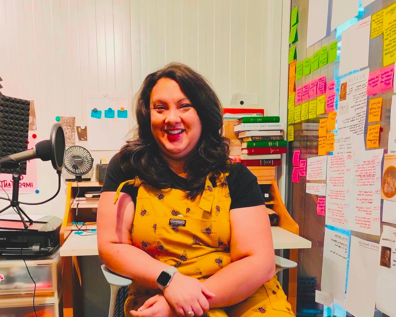 Author and branding strategist Amy Beeson sitting in her garden writing pod, smiling in a yellow dungaree outfit, surrounded by colourful post-its, storyboards, books, and a podcast mic—embodying the organised chaos of a writer mid-flow.