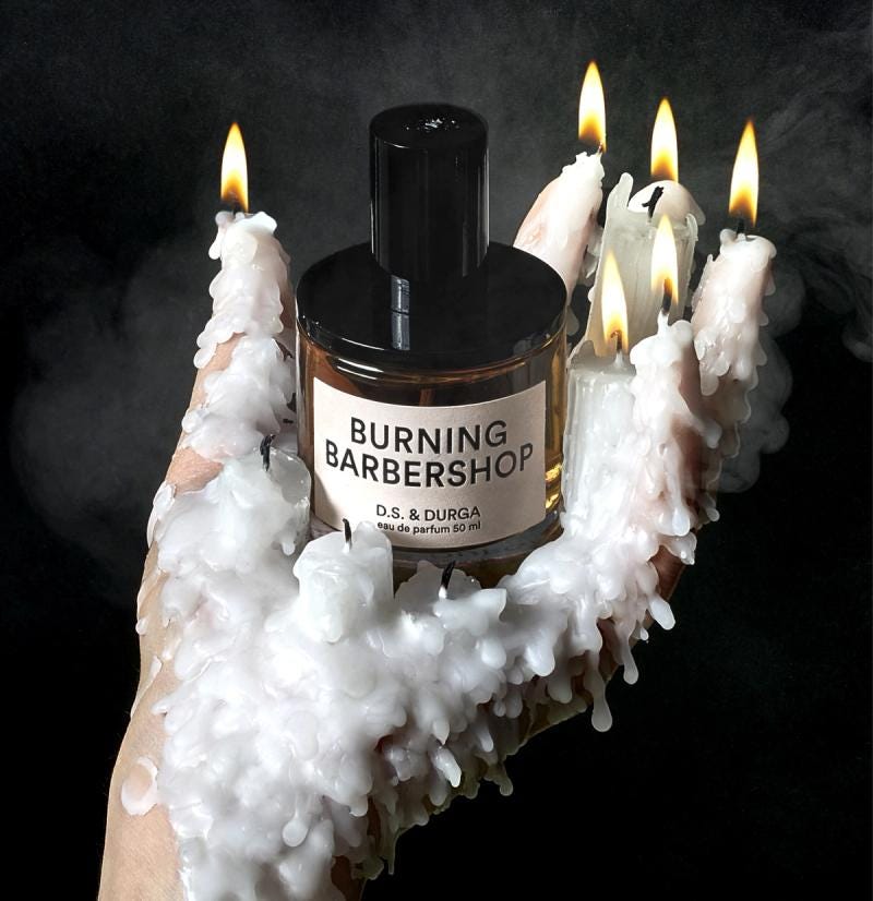 A bottle of Burning Barbershop fragrance by D.S. &amp; Durga is held by a hand covered in melted candle wax. Five lit candles are placed on the tips of the wax-covered fingers, producing small trails of smoke against a dark background.