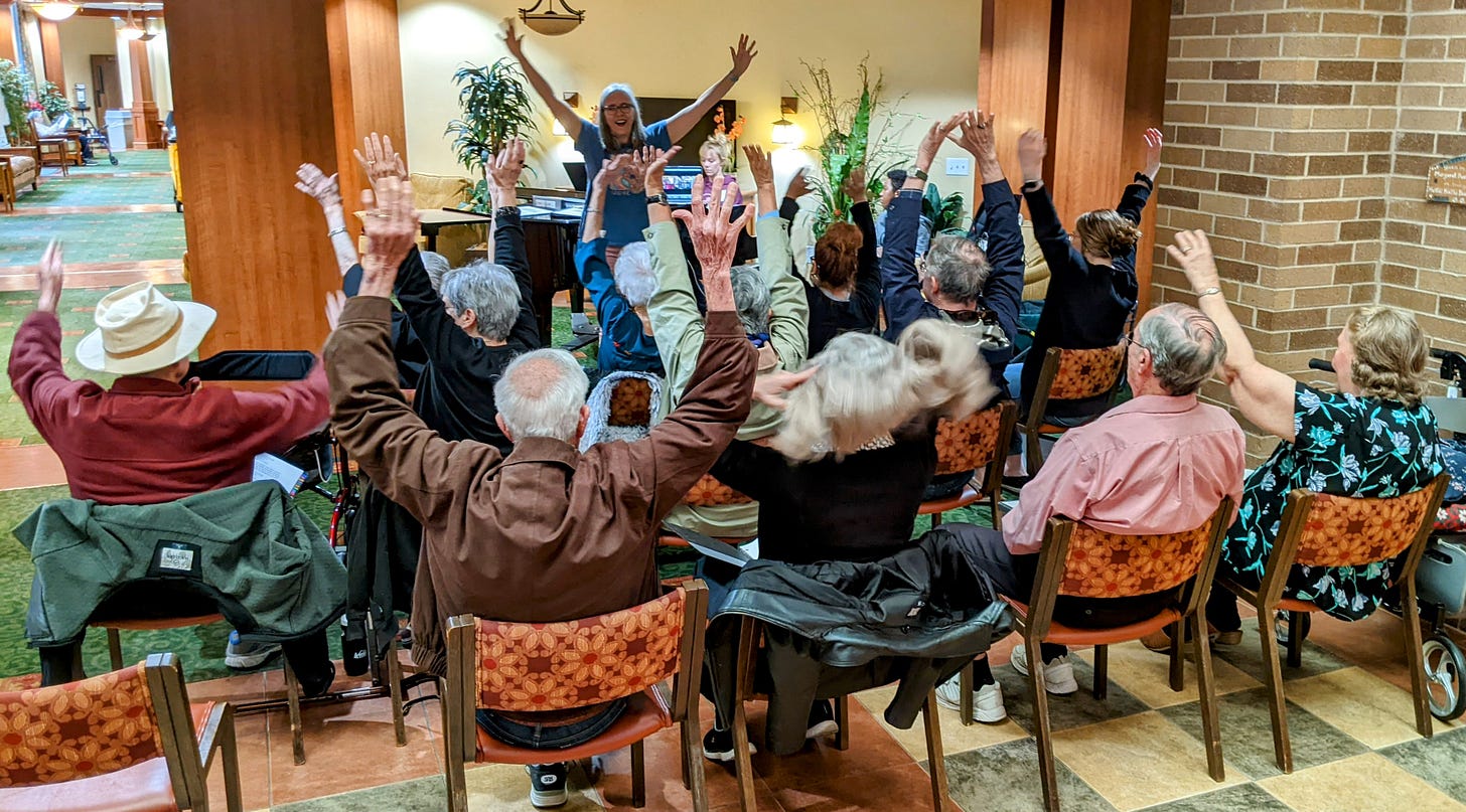 Silver-haired woman with arms lifted facing a group of senior adults with their arms raised.