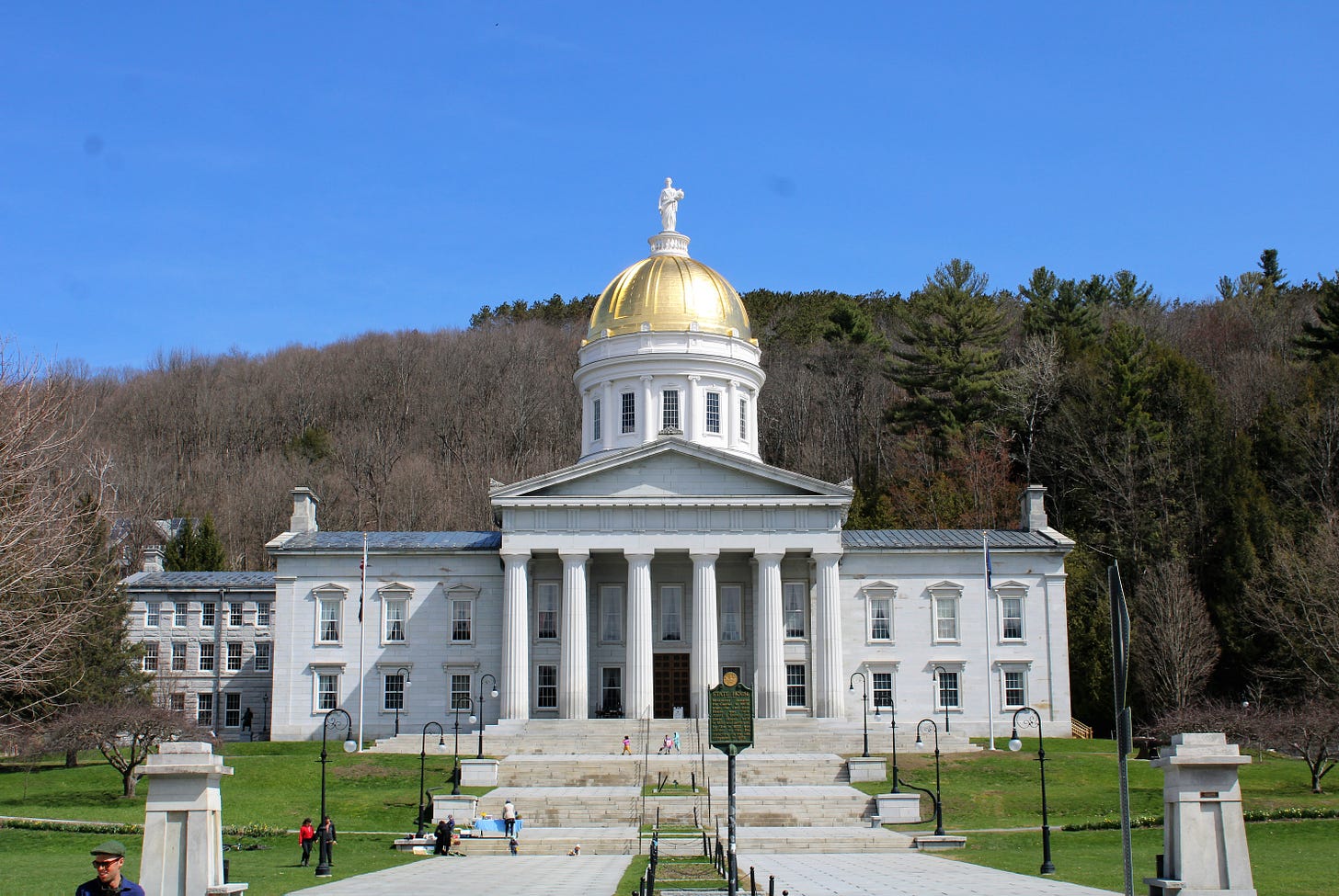 The Vermont State House in Montpelier, a white neoclassical building with tall columns and a gold-domed roof, set against a wooded hillside under a clear blue sky.