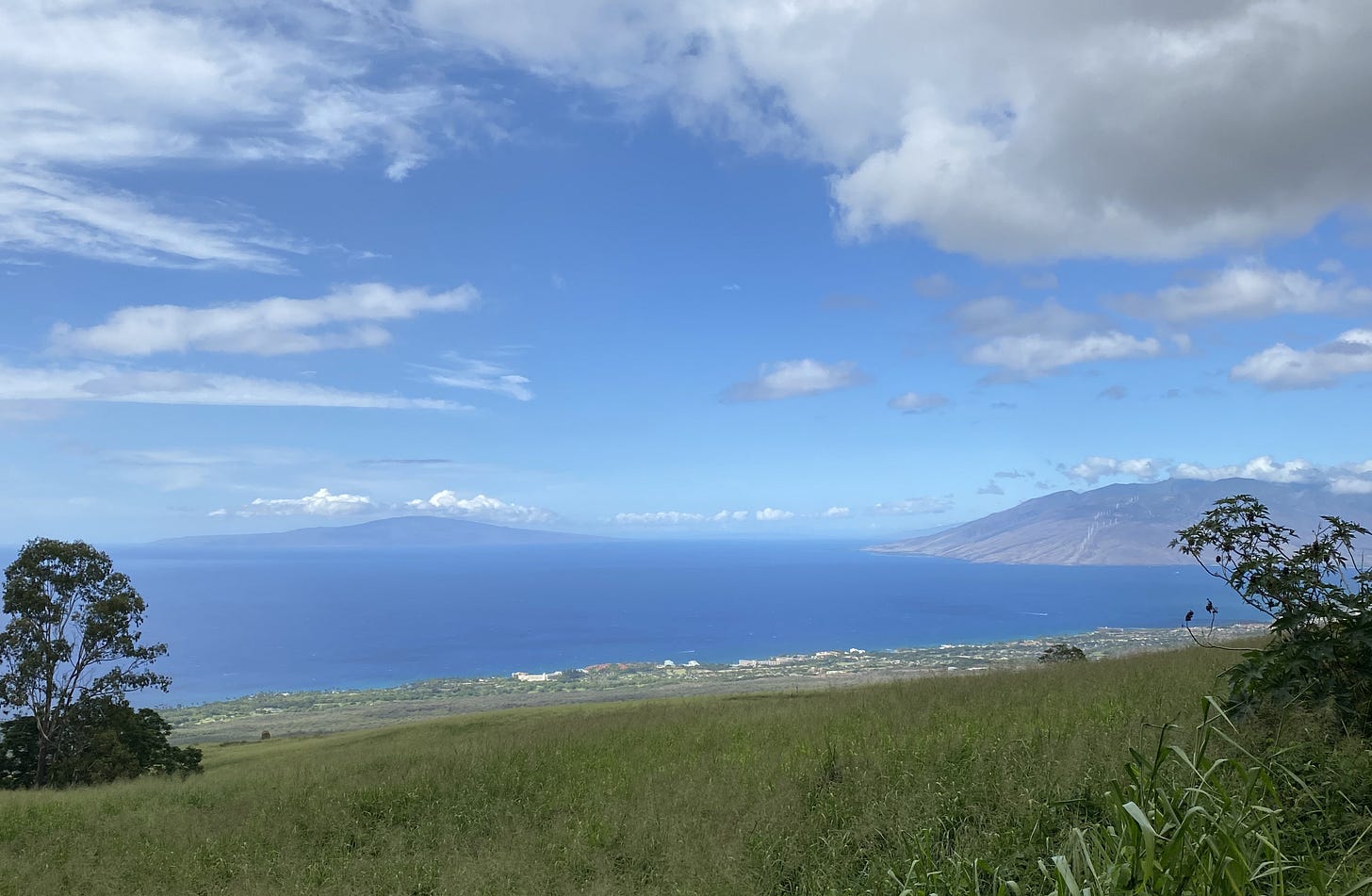 View over the Pacific from a small mountain road on South Maui. Gray clouds overhead, but in the distance the sky is vivid blue with a few puffy clouds here and there. The ocean under the blue sky is deep sapphire blue. At the right edge of the photo is another mountain, Maui’s second and older volcano, the West Mountain.