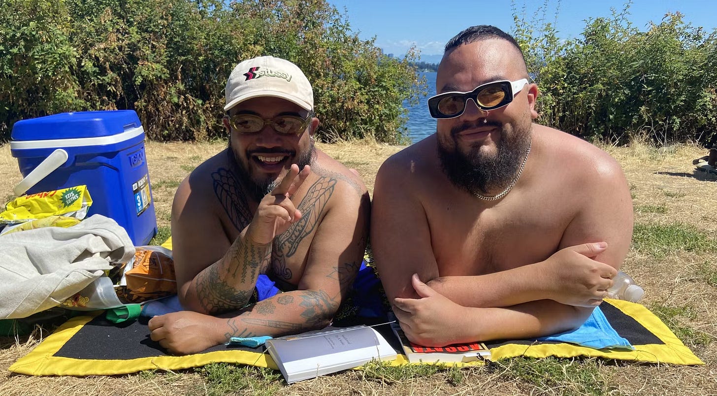 Two men, Derek Dizon and Erik Rios, sit together on a grassy area at Denny Blaine Park on July 15, 2025. They are relaxing in swimsuits on a sunny day, surrounded by greenery. Two men, Derek Dizon and Erik Rios, sit together on a grassy area at Denny Blaine Park on July 15, 2025. They are relaxing in swimsuits on a sunny day, surrounded by greenery.