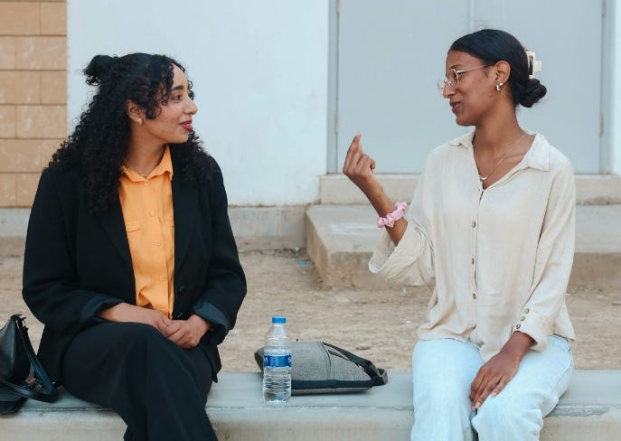 Two adult women of colour sit on a step outside having a discussion.