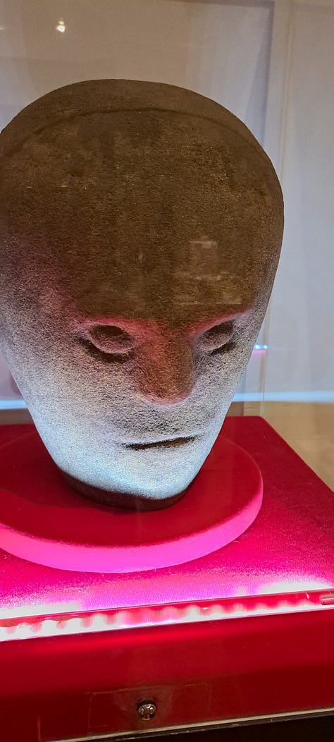 Two faces of the Corleck head on the top row with an image of a stamp featuring the Corleck head between them. Two images of other stone heads on display in our museum. All of them are made of a greyish-brown grainy stone, their facial features minimally represented and expressionless.
