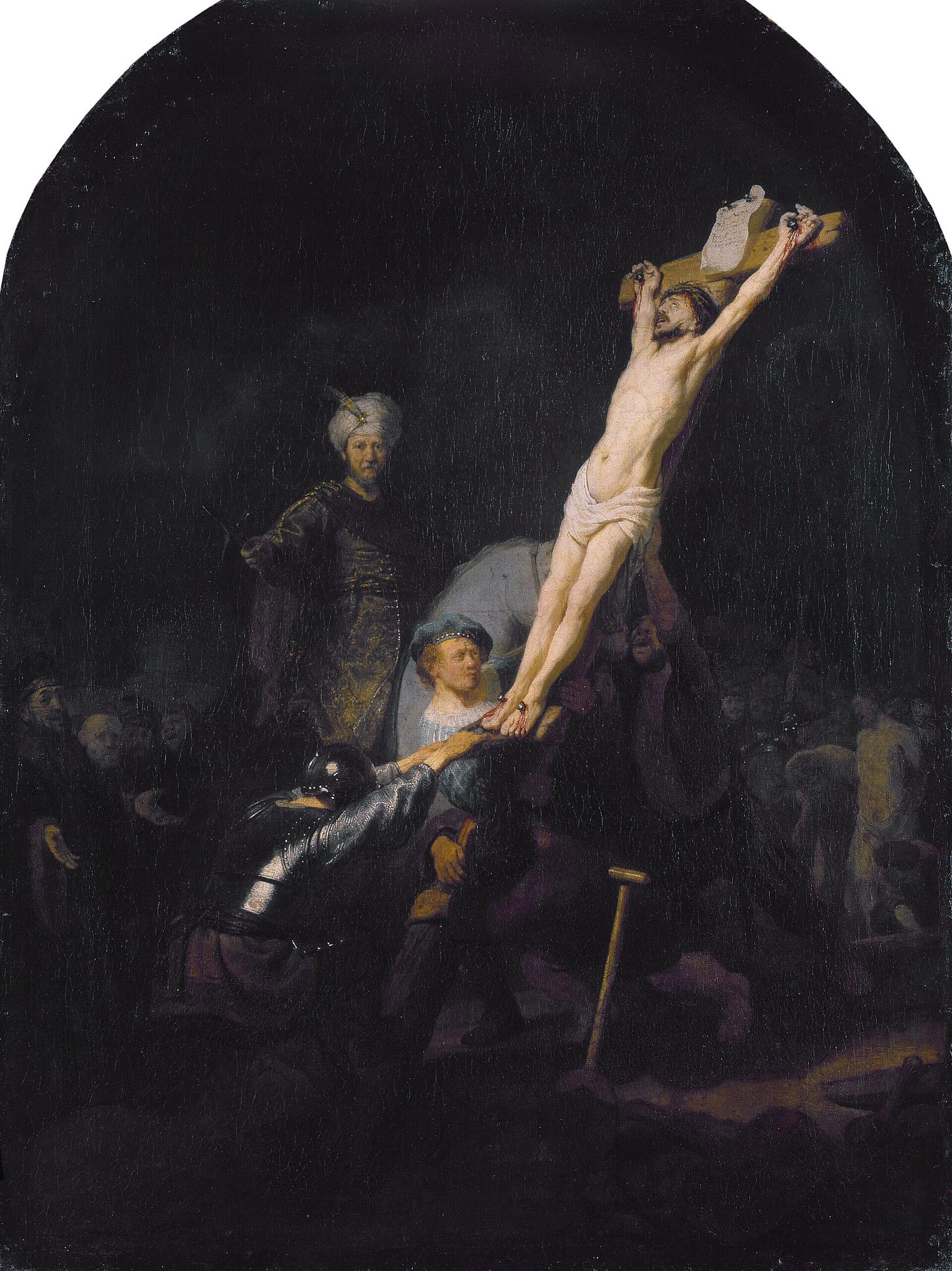 In a dark scene, men strain to haul upright a cross bearing Christ, whose pale body catches the light, one man in a blue beret grips the foot of the cross, and a horseman in a turban watches from the right. In a dark scene, men strain to haul upright a cross bearing Christ, whose pale body catches the light, one man in a blue beret grips the foot of the cross, and a horseman in a turban watches from the right.
