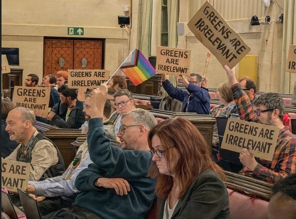 Bristol City Council chamber showing councillors holding 'GREENS ARE IRRELEVANT' placards and pride flags during public forum, illustrating the democratic crisis documented at https://bit.ly/4iCGBCh where elected representatives refused to listen to constituents