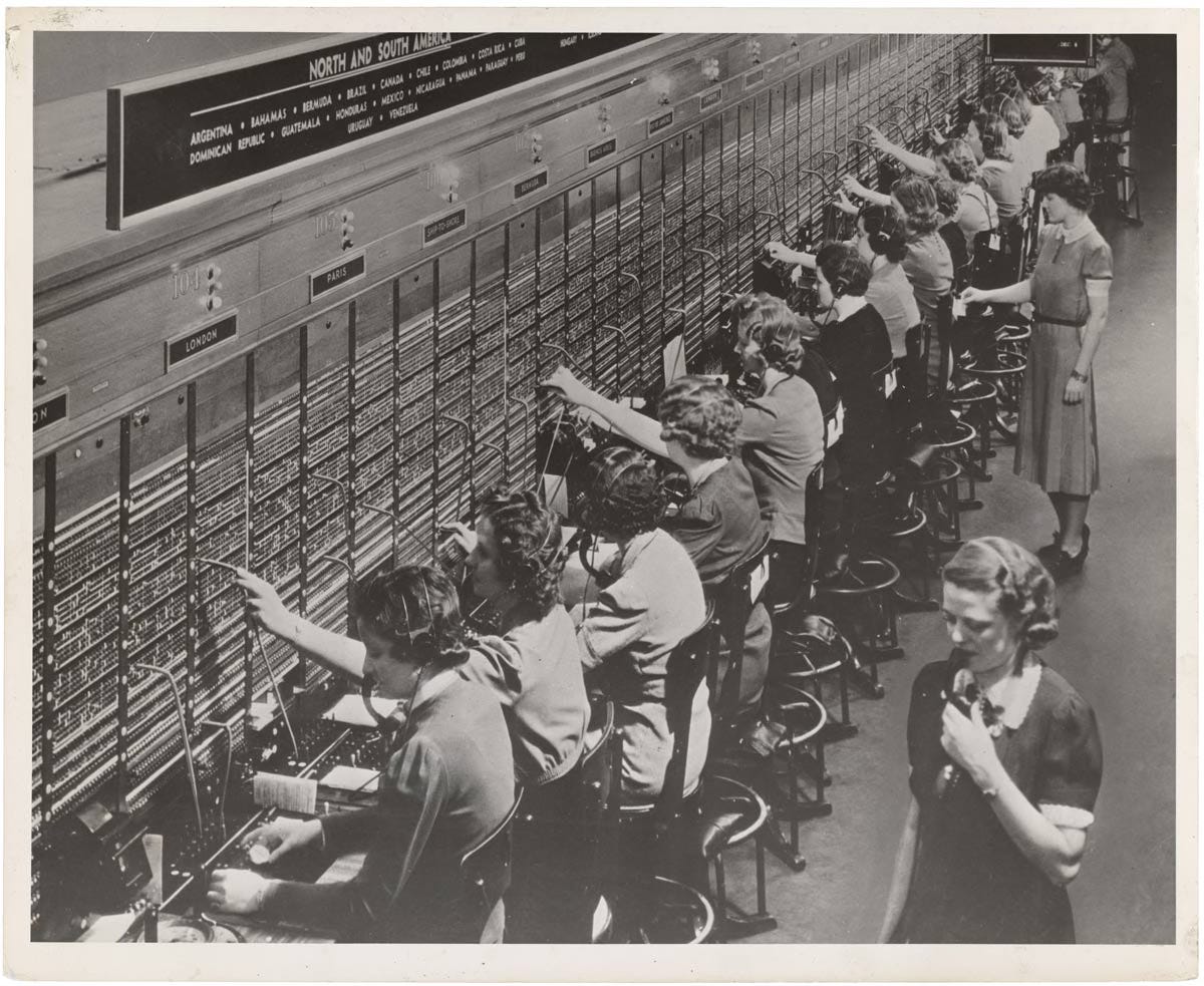 Black and white photograph of a large telephone switchboard operator room, circa mid-20th century. A long row of women wearing headsets sit at an expansive manual switchboard, inserting and pulling cables to connect international calls. A sign above reads "North and South America" with country names listed including Argentina, Brazil, Canada, and others. Labels on the board identify destinations such as London and Paris. A supervisor stands to the right observing the operators.