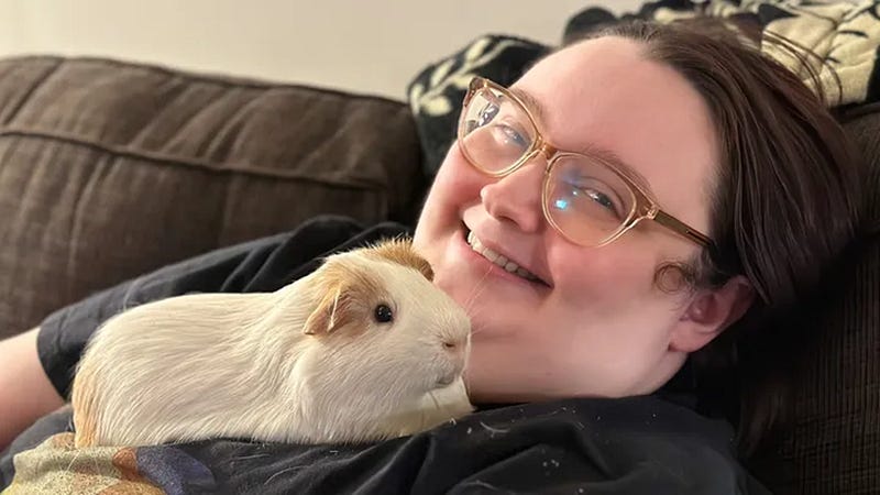 A smiling woman wearing glasses is looking at the camera and holding a white and tan guinea pig