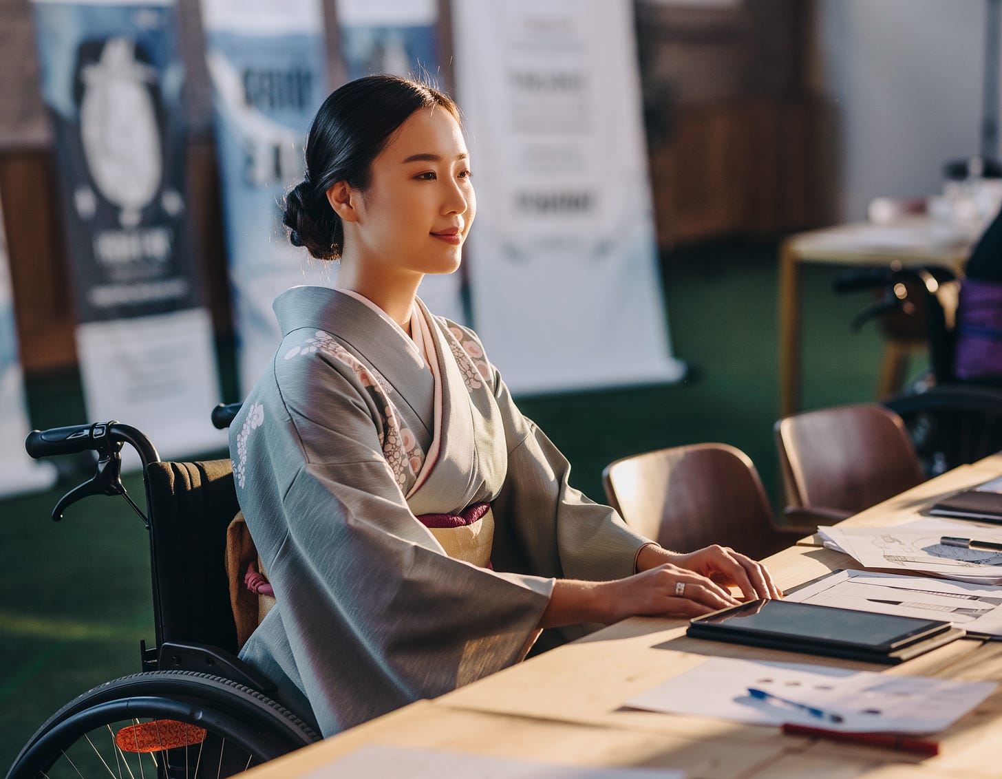 A young woman wearing an elegant gray kimono with delicate floral accents and a deep burgundy obi sits in a wheelchair at a long wooden table, her hand resting on a digital tablet as she gazes thoughtfully ahead. Her hair is styled in a neat updo, and warm golden sunlight bathes her in a soft, flattering glow. Scattered across the table are papers, sketches, and notebooks, suggesting she may be working on a creative or design project. In the background, roll-up banners and additional seating hint at a professional event, exhibition, or workshop setting, while another wheelchair is faintly visible in the far right, adding to the inclusive atmosphere of the space.