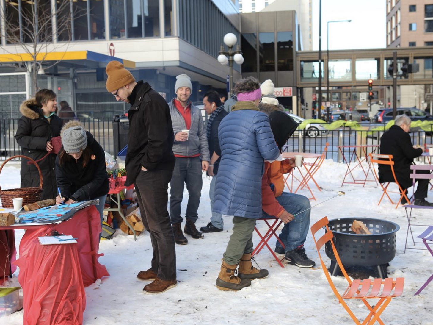 People gather around a snowy transit stop to roast s'mores and check out tables with information about the placemaking project happening there.