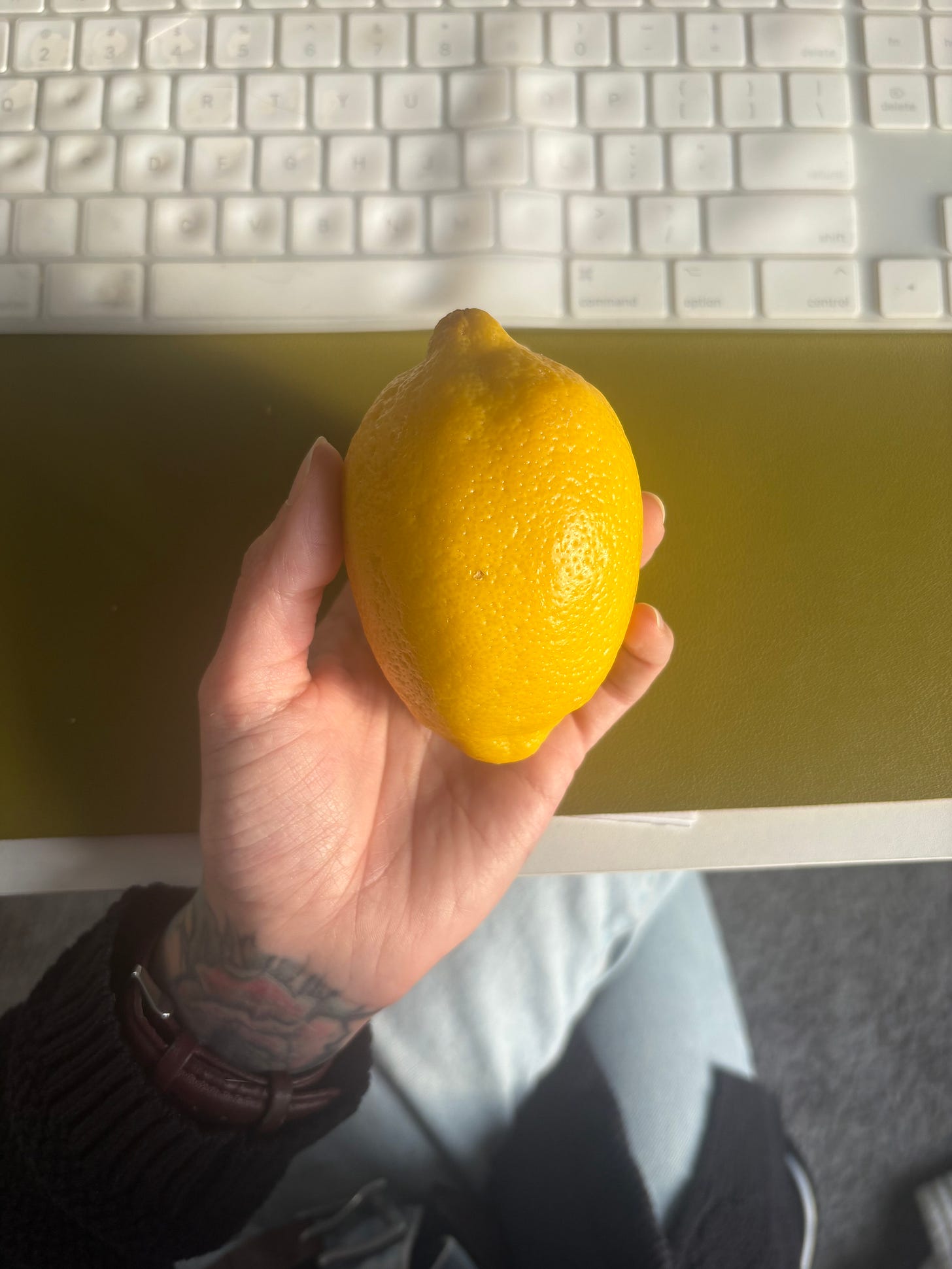 A hand with subtle tattoos holds a fresh yellow lemon above a desk, with a white keyboard softly blurred in the background. A hand with subtle tattoos holds a fresh yellow lemon above a desk, with a white keyboard softly blurred in the background.