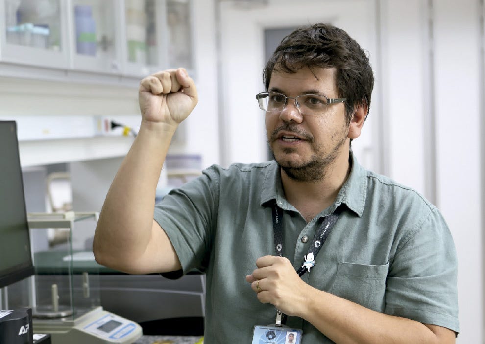 A imagem mostra um homem de pele clara, com cabelos curtos e castanhos, barba e bigode aparados, usando óculos de grau com armação metálica escura. Ele veste uma camisa de mangas curtas na cor verde-oliva e usa um crachá pendurado no pescoço por um cordão escuro. O homem está com o braço direito erguido e a mão fechada em um punho, enquanto sua mão esquerda está próxima ao peito, também parcialmente fechada, em um gesto que sugere uma explicação entusiasmada ou a demonstração de um conceito. Ele está em um laboratório, com o fundo levemente desfocado revelando bancadas brancas, armários com portas de vidro contendo frascos e o que parece ser o monitor de um computador à esquerda. A expressão facial é atenta e comunicativa, com a boca levemente aberta como se estivesse falando. A imagem mostra um homem de pele clara, com cabelos curtos e castanhos, barba e bigode aparados, usando óculos de grau com armação metálica escura. Ele veste uma camisa de mangas curtas na cor verde-oliva e usa um crachá pendurado no pescoço por um cordão escuro. O homem está com o braço direito erguido e a mão fechada em um punho, enquanto sua mão esquerda está próxima ao peito, também parcialmente fechada, em um gesto que sugere uma explicação entusiasmada ou a demonstração de um conceito. Ele está em um laboratório, com o fundo levemente desfocado revelando bancadas brancas, armários com portas de vidro contendo frascos e o que parece ser o monitor de um computador à esquerda. A expressão facial é atenta e comunicativa, com a boca levemente aberta como se estivesse falando.