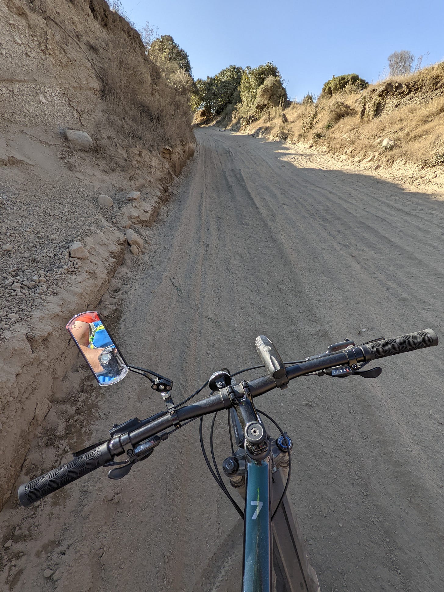 Handlebar view of a steep, dusty dirt road with ruts and loose gravel, surrounded by dry brush and low trees in rural Mexico.