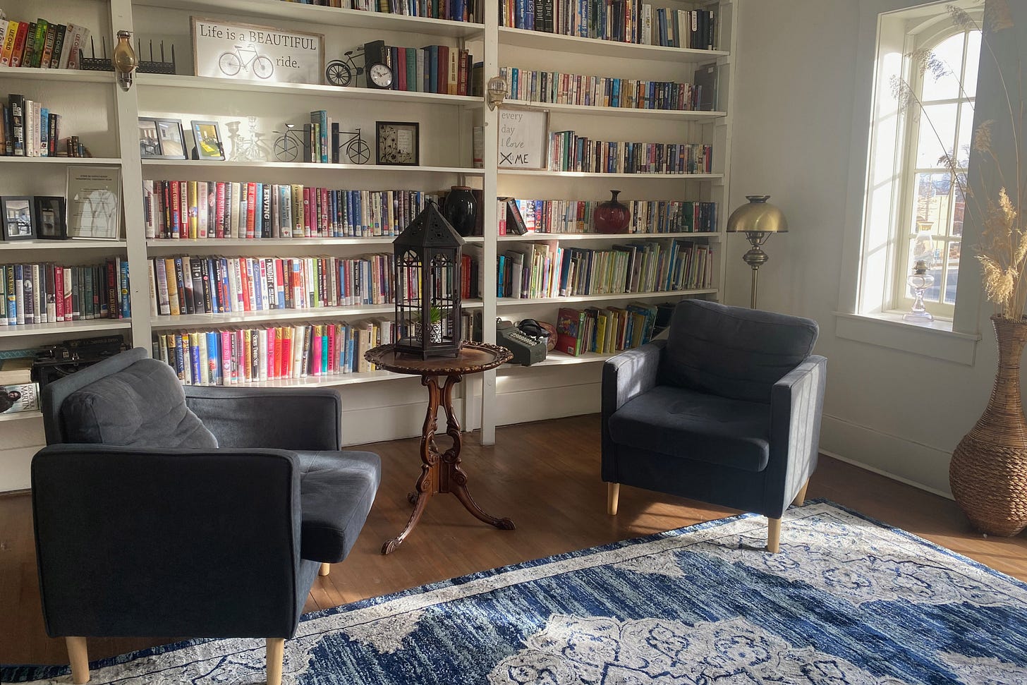 A room with two cushioned blue chairs in the center and a blue rug. The walls are lined with floor-to-ceiling bookshelves, filled with books with colorful spines. A window, on the right wall, lets in bright sunlight. A room with two cushioned blue chairs in the center and a blue rug. The walls are lined with floor-to-ceiling bookshelves, filled with books with colorful spines. A window, on the right wall, lets in bright sunlight.