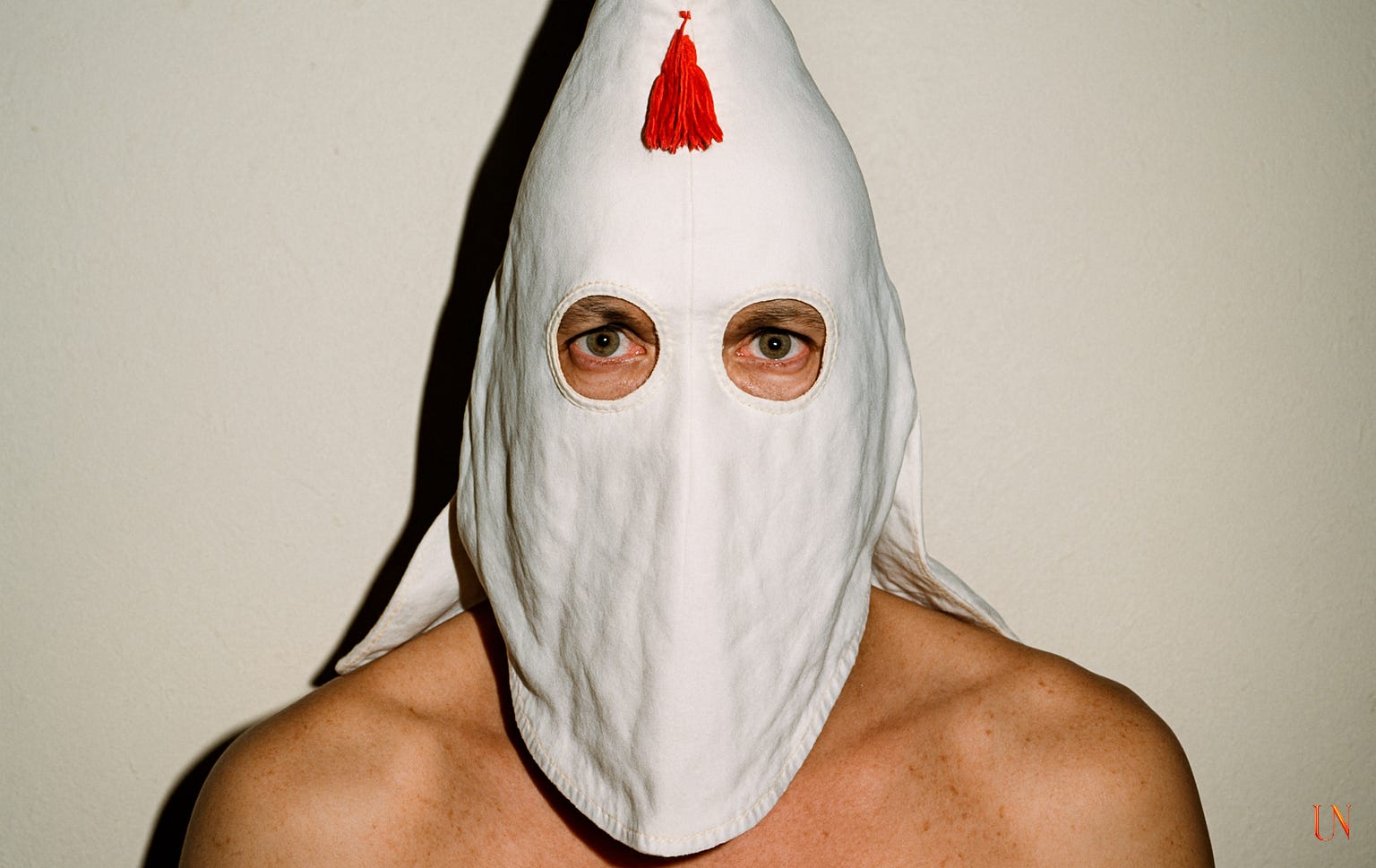 A stark color photograph of a man’s face covered by a white conical hood with stitched round eyeholes and a red tassel at the tip. The lighting is harsh and direct, flattening the image into near abstraction. His green eyes stare through the holes, expression calm but chilling. The background is pale and neutral, evoking the aesthetic of a forensic portrait.
