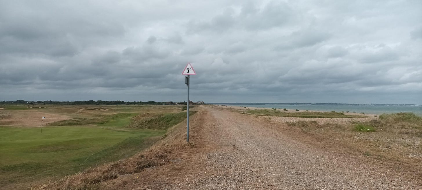 A path, on the left side is a golf course and on the right is the beach and the sea and in the middle is a sign with a person playing golf in a red warning triangle