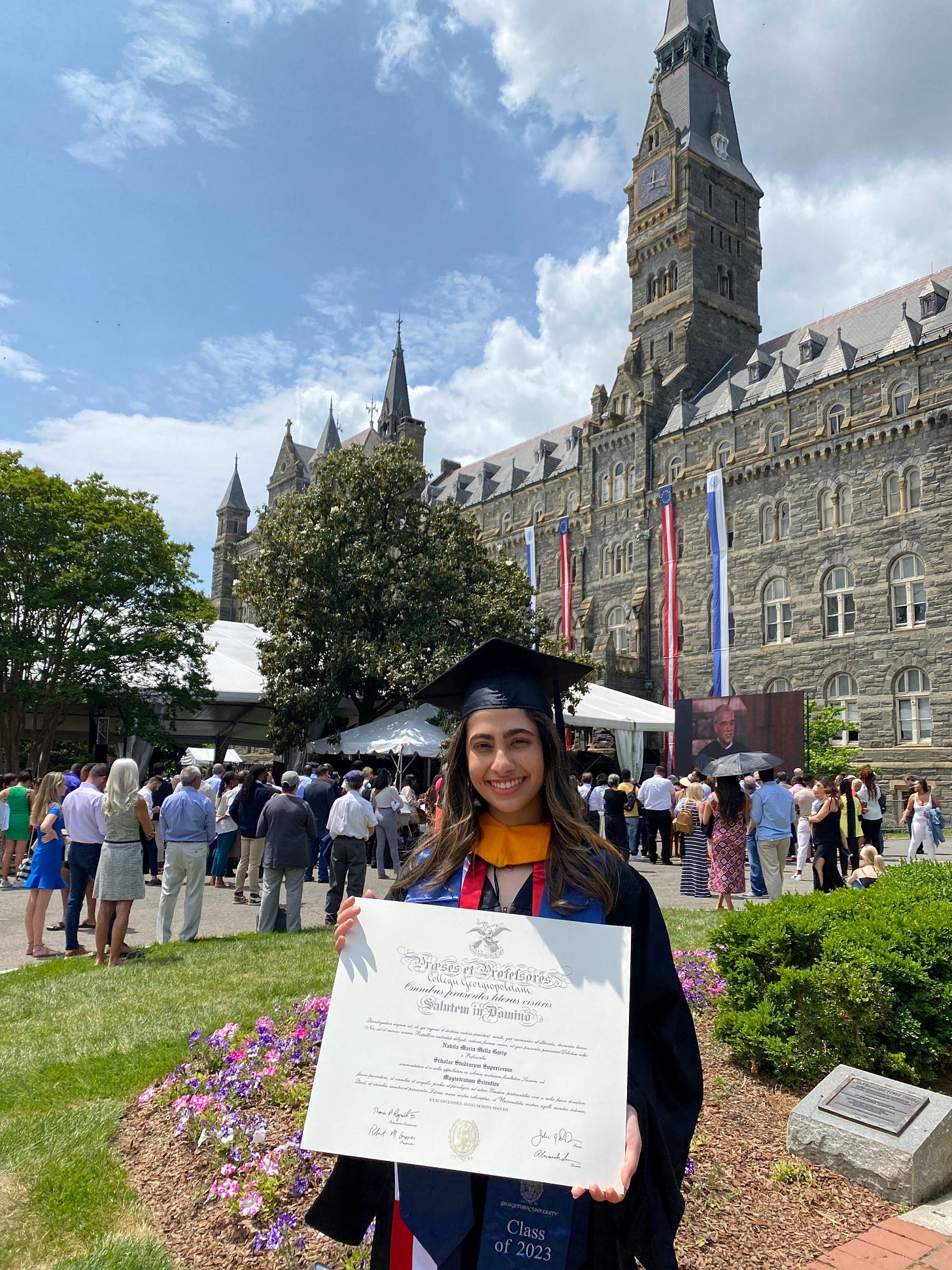 Nabila holding her Master's diploma at Georgetown University. It is sunny and she is smiling, wearing her cap and gown. 