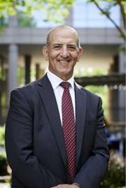 A bald man in a suit and red tie smiling in front of an office building