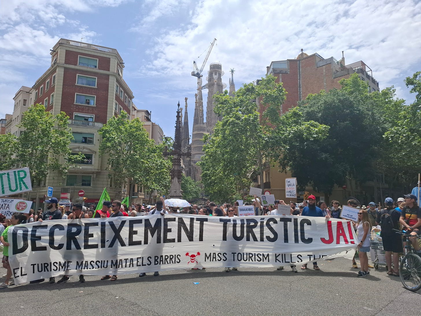 Una manifestazione di strada a Barcellona con un grande striscione orizzontale in primo piano che recita "DECREIXEMENT TURISTIC JA!!" (Decrescita turistica ora!!) in grandi lettere nere su sfondo bianco. Sotto questa frase, si legge "EL TURISME MASSIU MATA ELS BARRIS" (Il turismo di massa uccide i quartieri). Dietro lo striscione, una folla di persone partecipa alla manifestazione, alcune delle quali tengono altri cartelli. In lontananza, si possono vedere le guglie della Sagrada Familia.