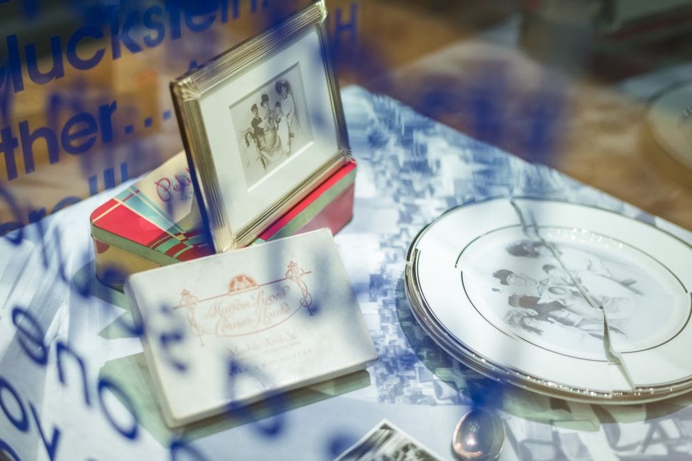 A glass display with blue text all over it contains a framed family photo, broken decorative plate, and an elegant card on a patterned tablecloth. A glass display with blue text all over it contains a framed family photo, broken decorative plate, and an elegant card on a patterned tablecloth.