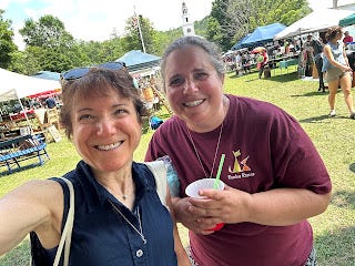 Me and my sister, smiling, with tents and vendors in the background across a green lawn. Me and my sister, smiling, with tents and vendors in the background across a green lawn.