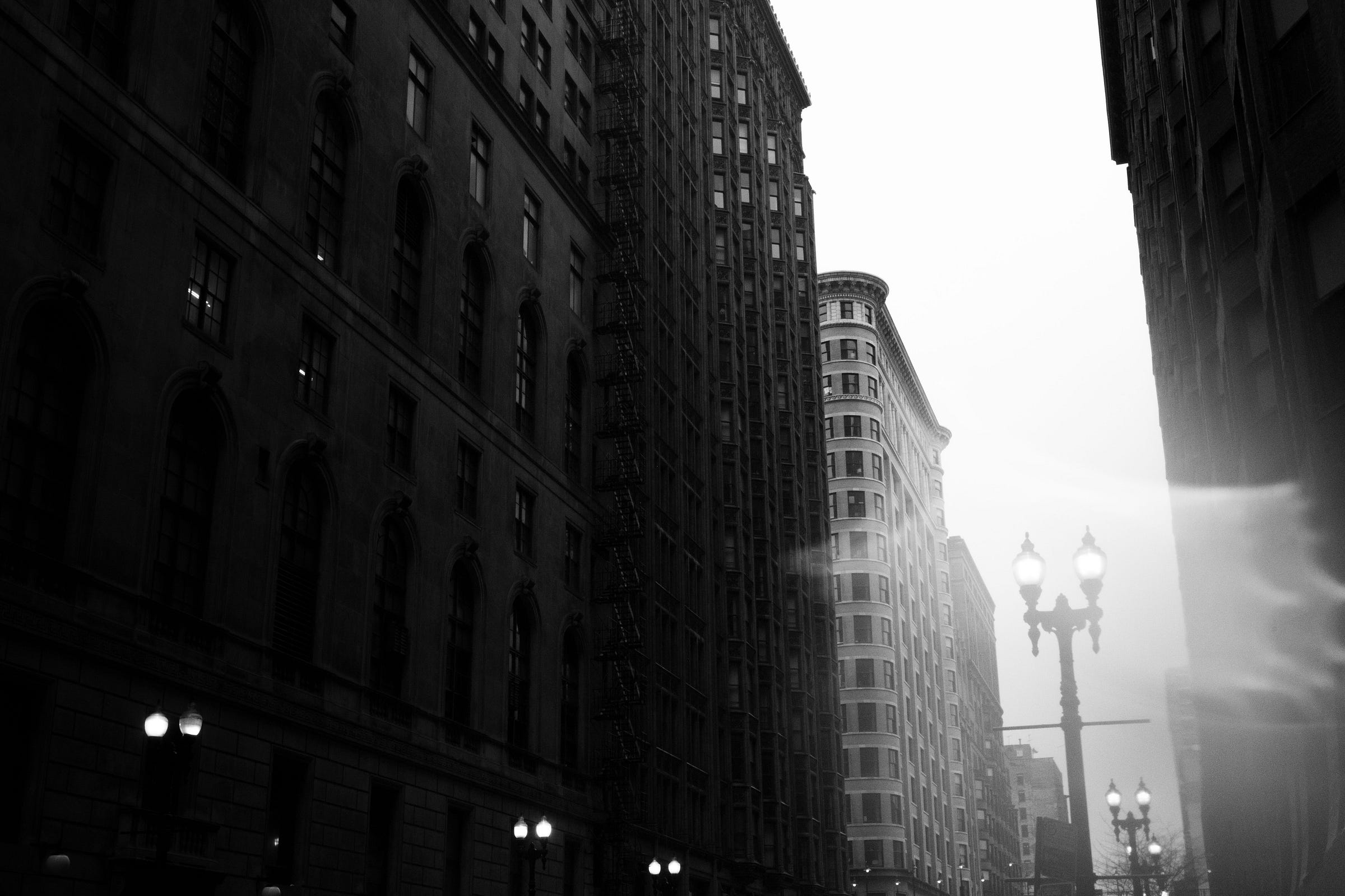 A moody black-and-white cityscape featuring towering historic buildings with glowing streetlights and misty light streaks in the foreground, creating a dramatic atmosphere. A moody black-and-white cityscape featuring towering historic buildings with glowing streetlights and misty light streaks in the foreground, creating a dramatic atmosphere.