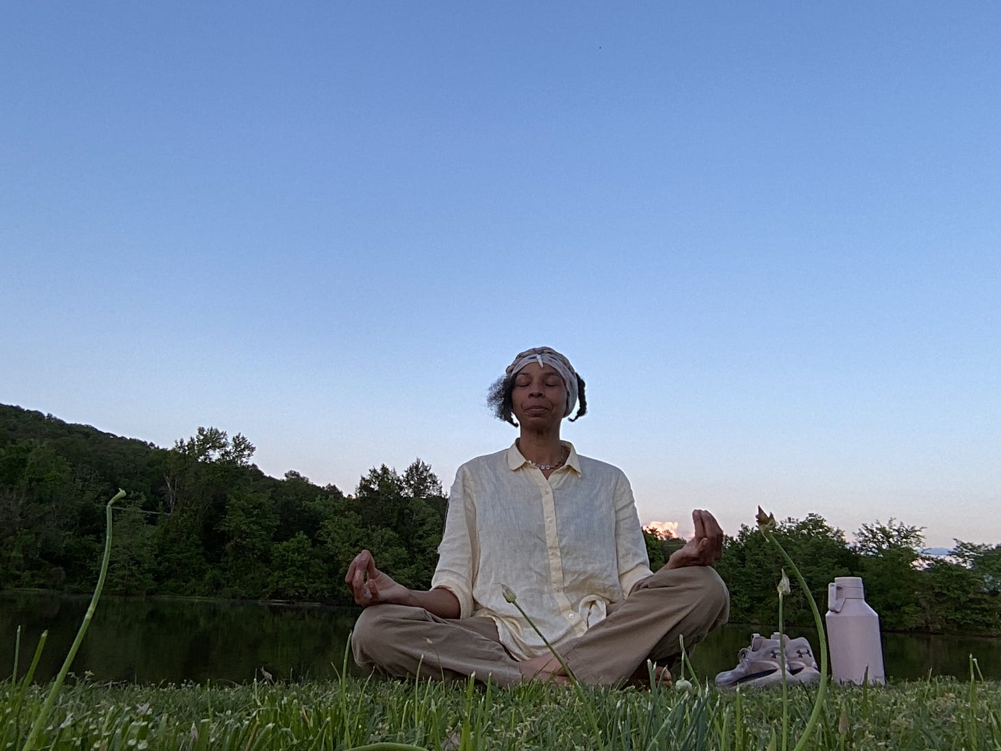 Lanita sitting in meditation mode eyes closed, finger tips touching with elbows rested on knees, next to a lake with a forest background and clear sunset skies. Pink sneakers and water bottle resting next to her in the grass. 
