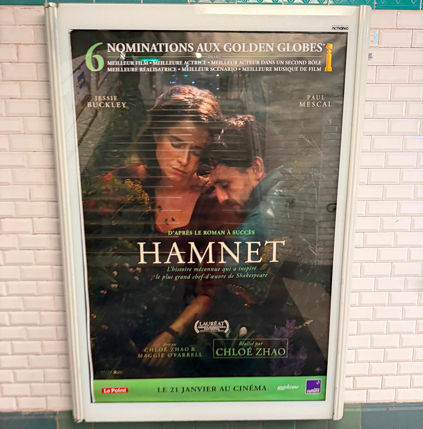 Movie poster for *Hamnet*, directed by Chloé Zhao, displayed in a Paris Métro station with white tile walls and green Art Nouveau trim. The poster shows Jessie Buckley and Paul Mescal in a close embrace surrounded by greenery. Text in French reads "D'après le roman à succès" and "L'histoire méconnue qui a inspiré le plus grand chef-d'œuvre de Shakespeare." A banner at the top highlights 6 Golden Globe nominations. Release date: January 21. Movie poster for *Hamnet*, directed by Chloé Zhao, displayed in a Paris Métro station with white tile walls and green Art Nouveau trim. The poster shows Jessie Buckley and Paul Mescal in a close embrace surrounded by greenery. Text in French reads "D'après le roman à succès" and "L'histoire méconnue qui a inspiré le plus grand chef-d'œuvre de Shakespeare." A banner at the top highlights 6 Golden Globe nominations. Release date: January 21.