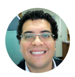 Homem branco, cabelos e olhos castanhos, usa óculos, camisa branca e blusa preta. Esboça leve sorriso