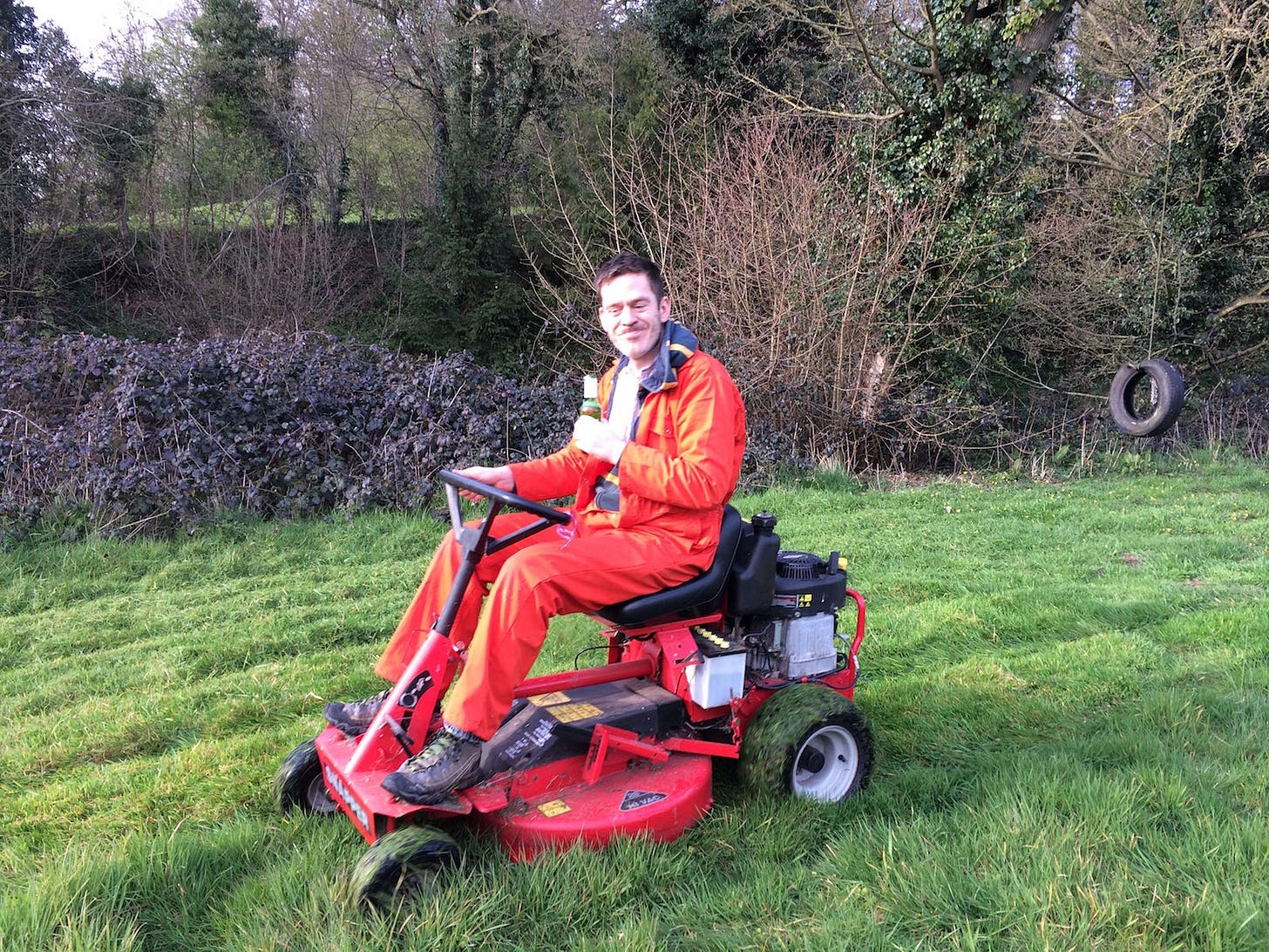 Joe is a white man wearing an orange jumpsuit and a huge grin, holding a bottle of beer and riding a red motor mower on an unkempt lawn, brambles in the background