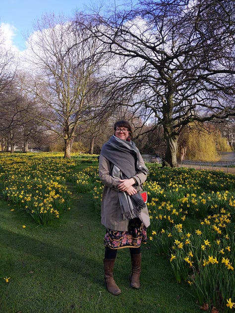 Three photos of me in St. James' Park in February 2020. I am standing surrounded by daffodils, and whilst I am rugged up in my grey coat and blanket scarf, a floral skirt peeks out from underneath and I am clearly embracing the new season. The background includes three big tree still in Winter mode but a small patch of blue sky is peeking through the clouds. Photos taken by S-
