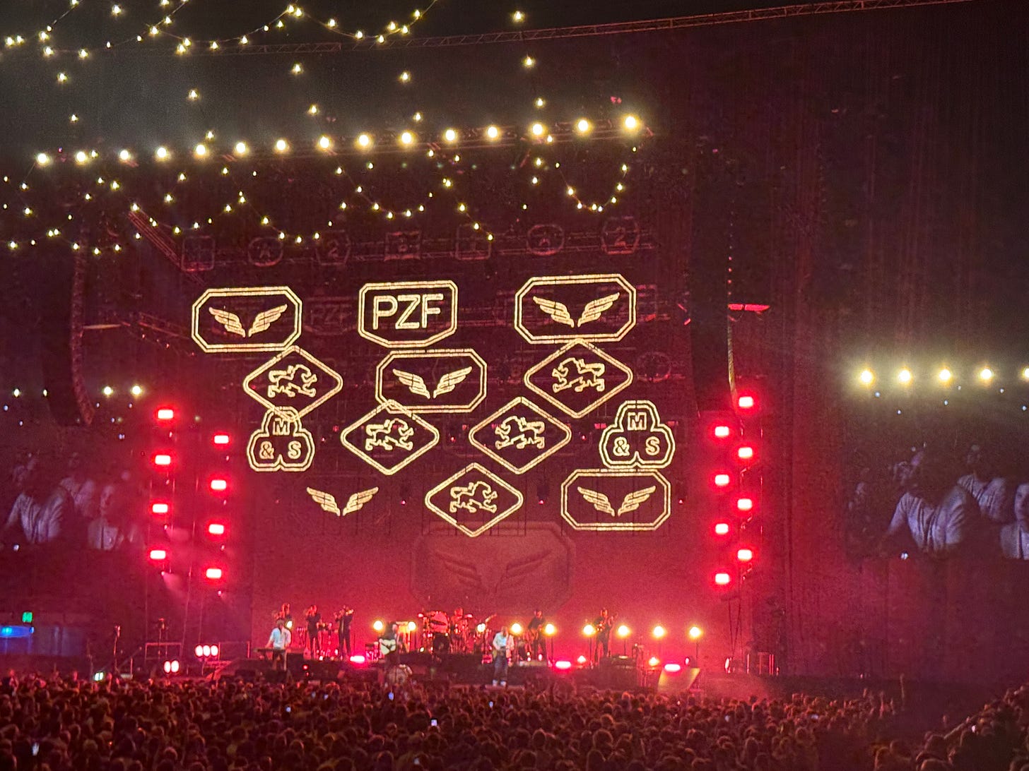 Photo of the staging of the Mumford and Sons concert, Various images in lights and string lines giving the room a barn vibe. Red lighting in the background.