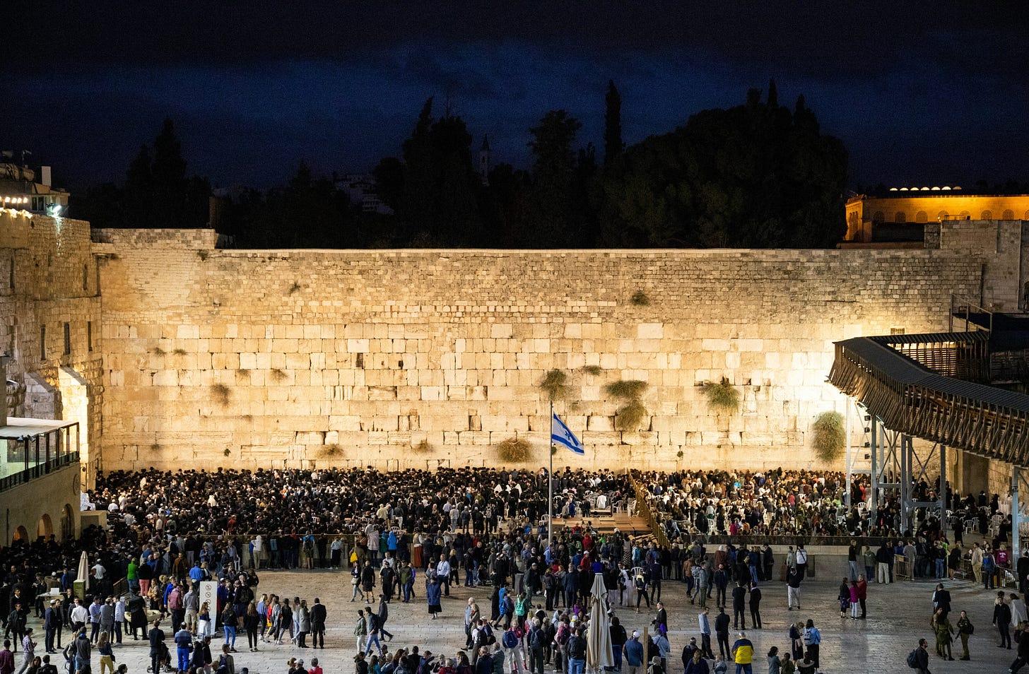 Photo of a crowd at the Western Wall at night, blue-black sky and golden lights on the wall