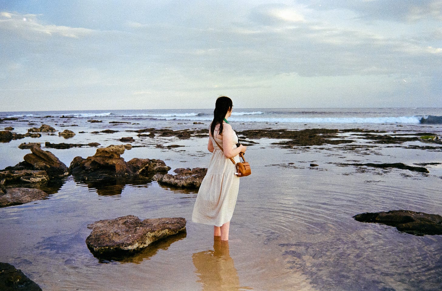 A photo of Josie in a flowing beige dress with her brown leather handbag and camera, feet in the ocean up to her ankle, as the breeze softly blows her dress, hair, and creates ripples all round her. A photo of Josie in a flowing beige dress with her brown leather handbag and camera, feet in the ocean up to her ankle, as the breeze softly blows her dress, hair, and creates ripples all round her.