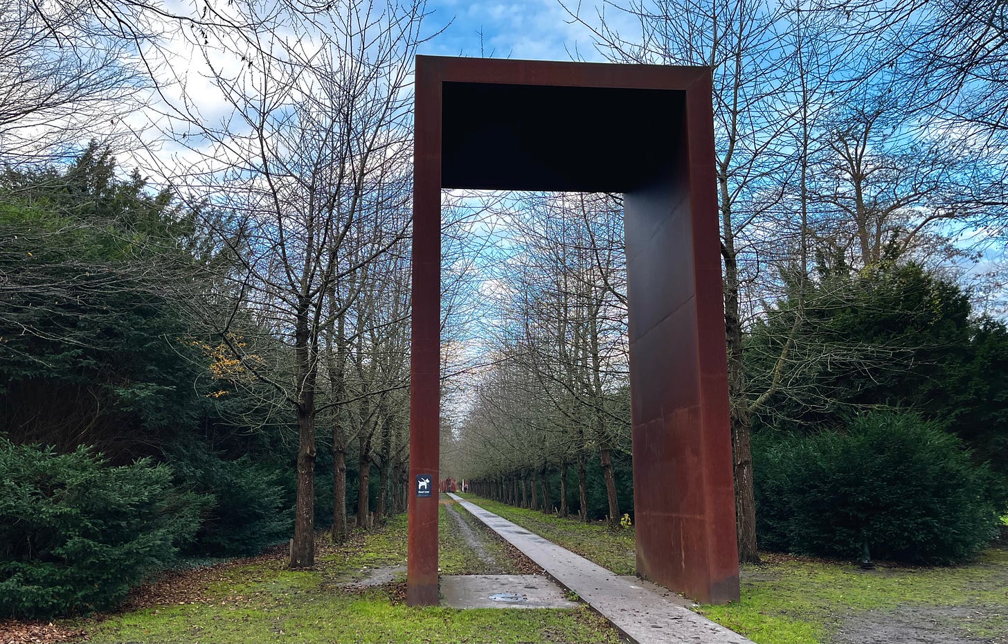 squared, contemporary, rusted iron arch over a straight path leading into the distance between high evergreen shrubs