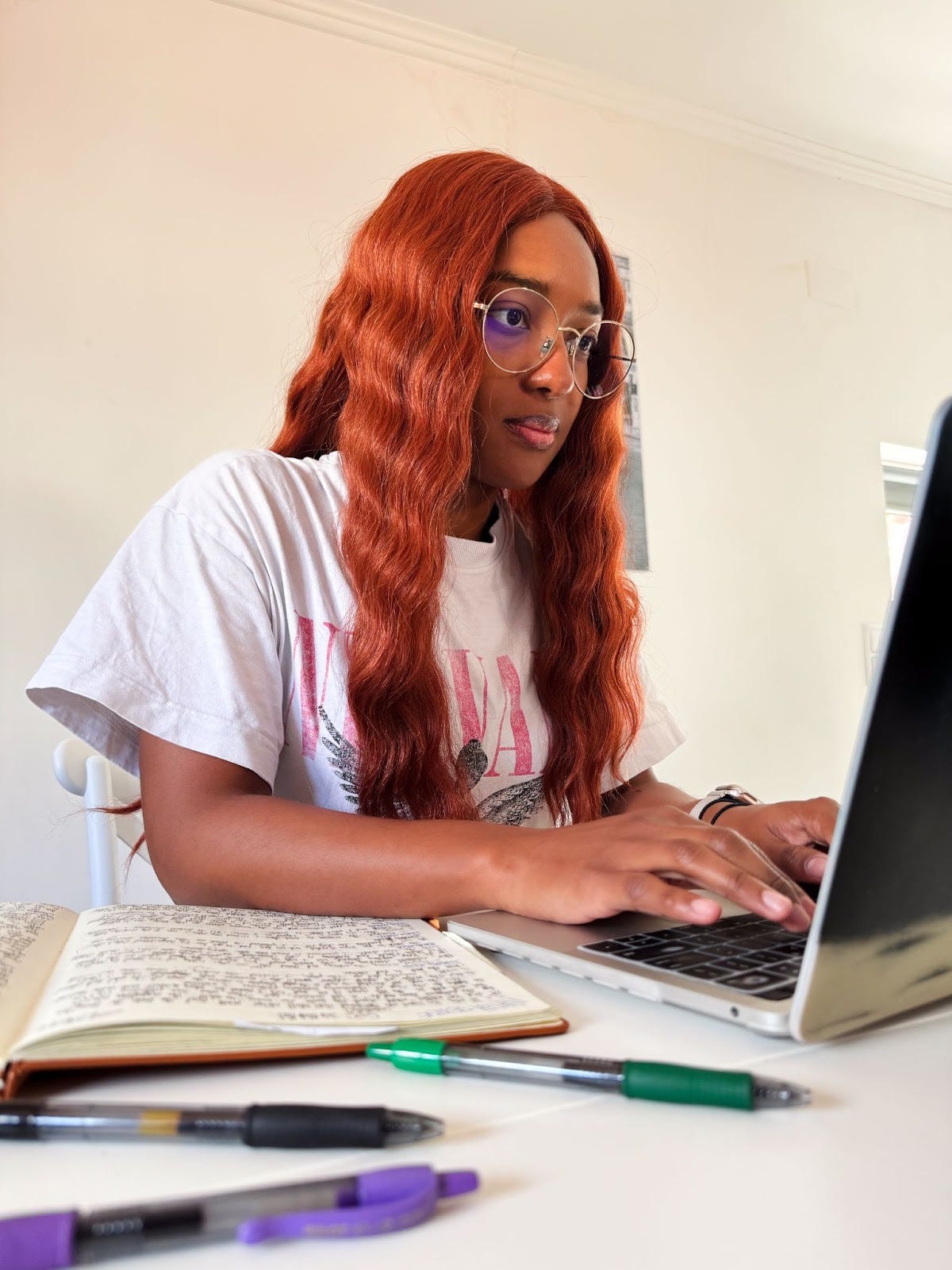 A photo of Kat Lewis, a Black woman with curly, warm-colored hair. She types on her laptop with a notebook opened on her desk. A photo of Kat Lewis, a Black woman with curly, warm-colored hair. She types on her laptop with a notebook opened on her desk.
