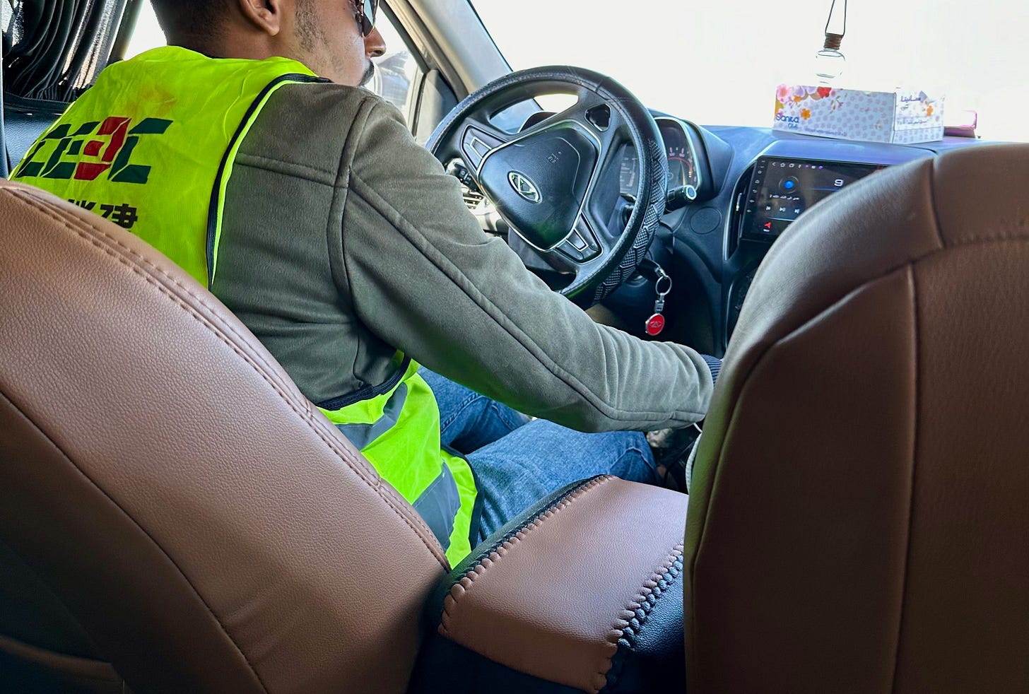 A driver seen from the back seat wearing a neon yellow high-visibility vest with Chinese characters from China Energy Engineering Corporation, gripping the steering wheel with a strawberry air freshener hanging from the rearview mirror
