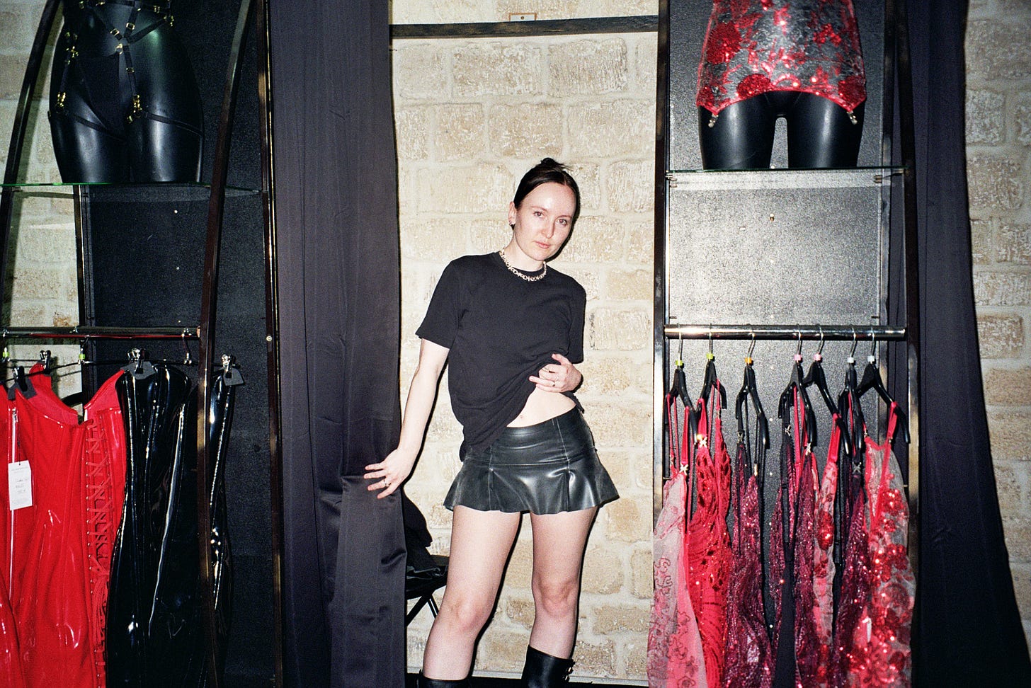 Evyn Silver, a white femme with dark hair worn in a French twist, poses while pulling back a dressing room curtain in a stone basement. With the other hand, she lifts her black t-shirt to show off a black latex mini skirt.