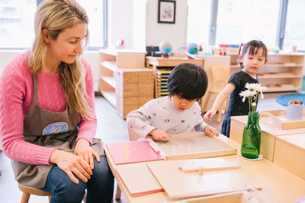Montessori guide observing a child tracing a sand tray for letter formation — what is Montessori teacher role
