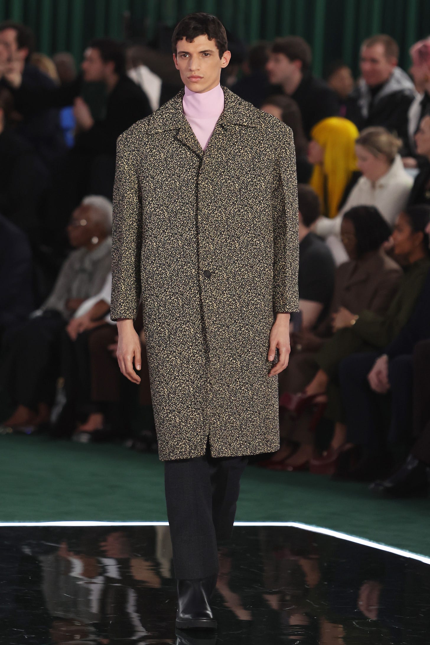 A male model walks a runway wearing a strictly tailored grey melange cloth coat over a pale pink turtelneck with black pants.
