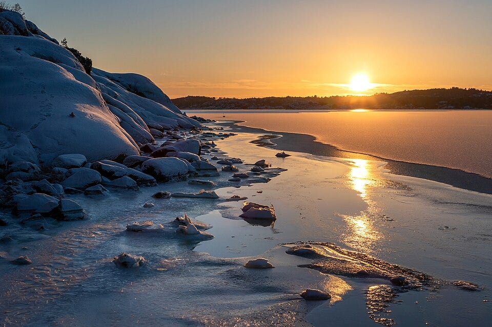 Golden setting sunlight on water, coast, and snowy, rocky hills.