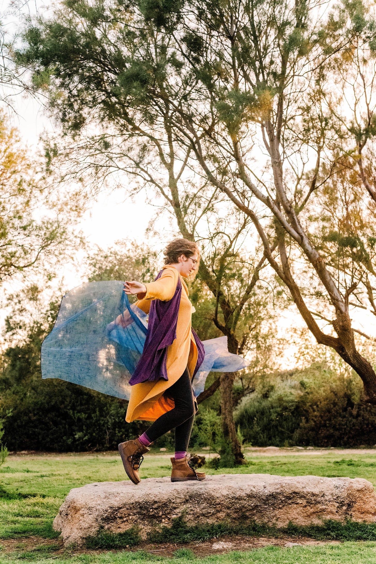 Photo of the author wearing a golden dress, black leggings, and purple vest dancing with fabric on a rock between trees in the golden hour