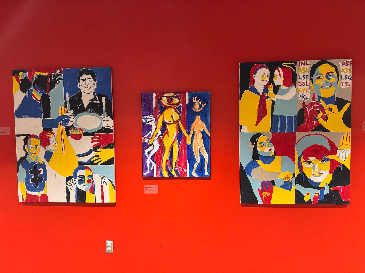 A bright red wall filled with three paintings by Nancy Rourke. All are in her trademark red-blue-yellow bold style and feature deaf people and motifs.