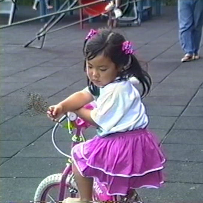 Photo of a girl with pig tails wearing pink bows and pink skirt, contemplating a branch in her hand as she sits on a pink tricycle.