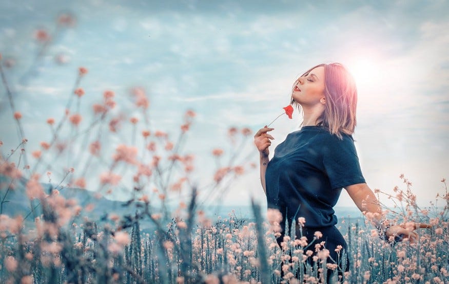 Woman standing in a field of flowers with her eyes closed holding a red poppy under her chin
