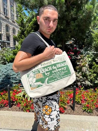 Man carrying a canvas New York Transit Museum tote printed with a vintage B.M.T. advertisement reading “Reach the Beach by Subway.” Man carrying a canvas New York Transit Museum tote printed with a vintage B.M.T. advertisement reading “Reach the Beach by Subway.”