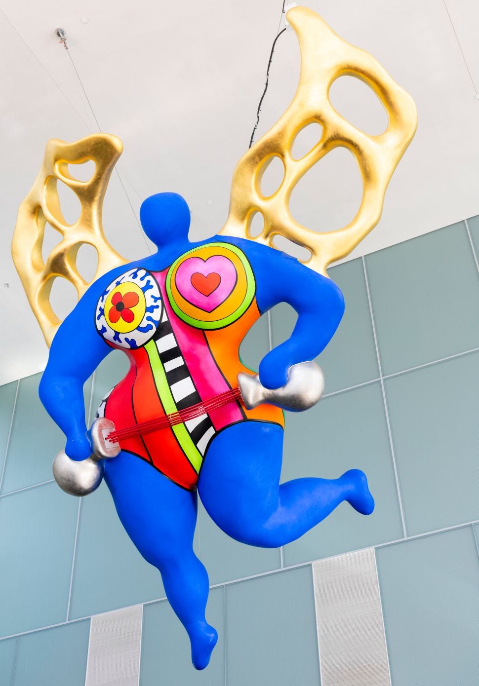 A colorful sculpture of a blue, winged female figure holding weights stands in front of a modern architectural wall, with its wings designed in a golden lattice pattern.