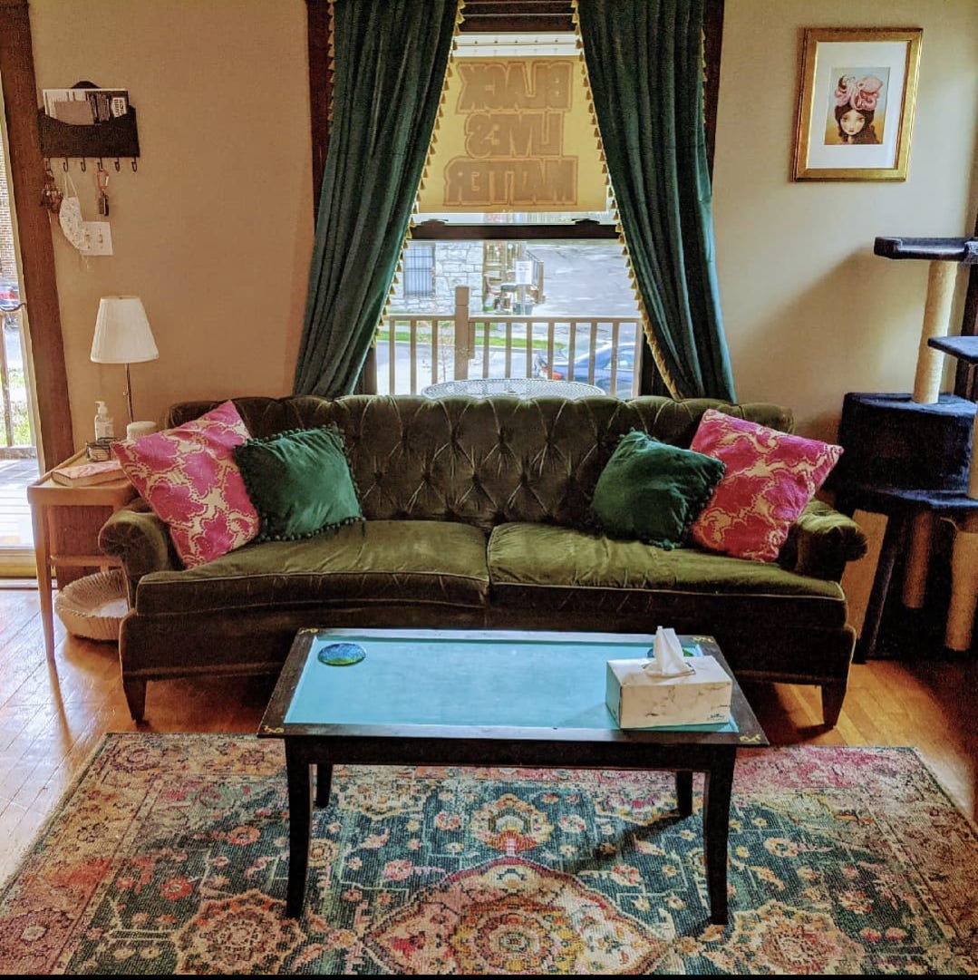 A photograph of a brightly lit living room. The centerpiece is a tufted green velvet sofa underneath a picture window, framed with heavy turquoise curtains. The sofa has green and pink throw pillows. There is a lamp to the left of the sofa and a cat tower to the right. In front of the sofa, a black and turquoise coffee table sits on top of a large rug with intricate designs in pink, green, turquoise, and cream.