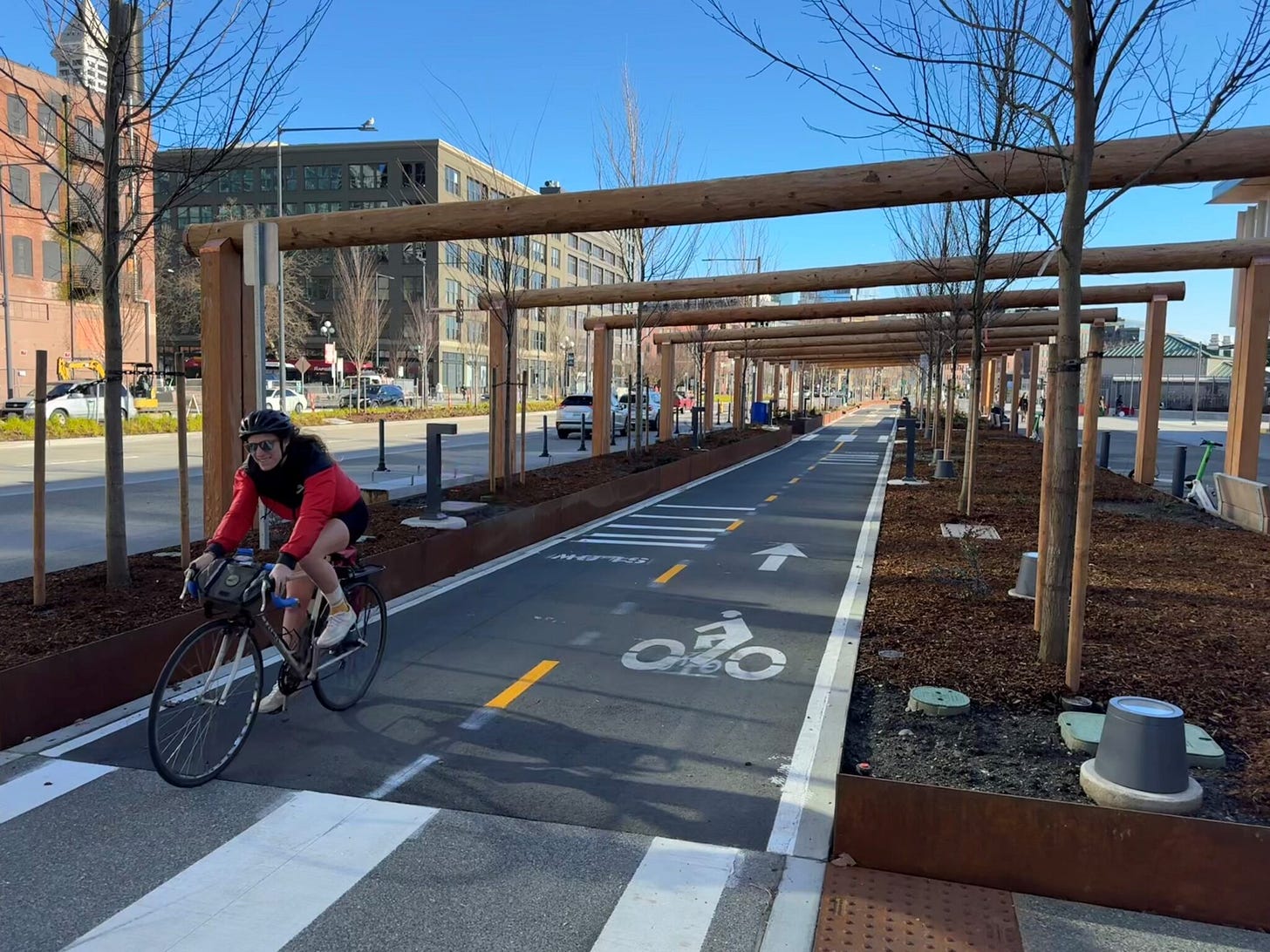 A person wearing shorts and a red and black jacket and helmet bikes along a bike lane. Buildings and blue skies are in the background, along with landscaped woodchips in planter areas next to the bike lane.